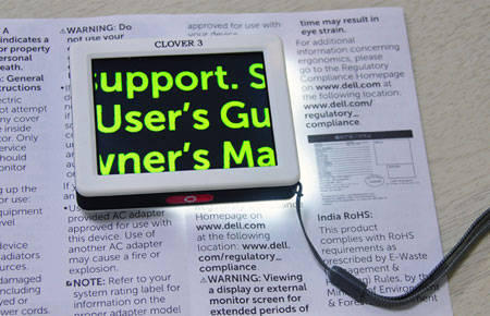 Clover 3 pocket beeldschermloepje op een blad tekst. Op het schermpje is de tekst vergroot te zien; gele letters tegen een zwarte achtergrond.