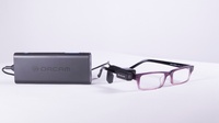 Een bril waarop de camera vastgeklipt is en waaruit een draad loopt naar het besturingskastje dat voor spraakuitvoer zorgt.