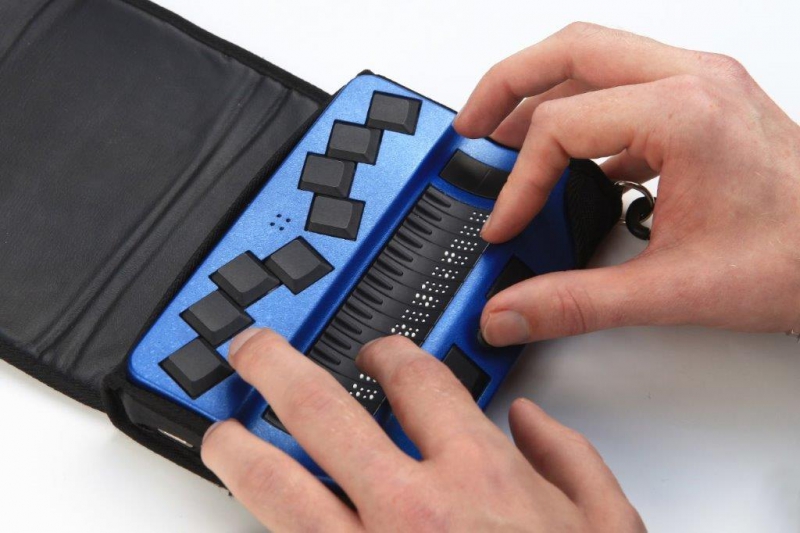 Blauw apparaatje met brailletoetsenbord en 16 braillecellen. Het bevindt zich in een zwart etui. Twee handen voelen aan de braillecellen en de functietoetsen op de voorzijde.