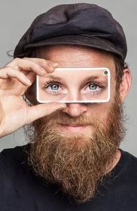 Man met baard en zwarte pet die een smartphone voor zijn ogen houdt. Op de smartphone zijn 2 vrouwenogen te zien.