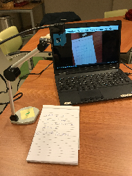 De Scan2Voice camera in uitvouwen toestand. De camera staat gericht op een tekst op papier op de tafel, de  tekst wordt geprojecteerd op een laptop die ernaast staat.