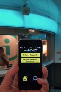  Foto van een smartphone waarop de myMOVEO app werkt met op de achtergrond een loket waarboven een sprekend baken geplaatst is.